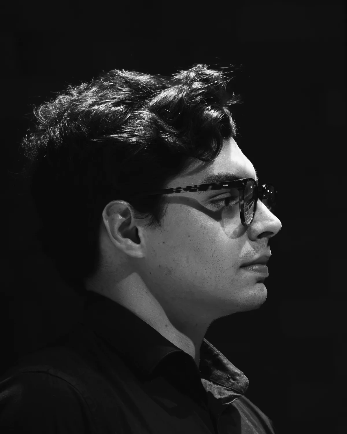 Retrato em preto e branco de Lucas Quaranta de perfil, do peito para cima, sobre fundo preto. A luz lateral desenha o cabelo ondulado, a armação dos óculos e a linha do maxilar.