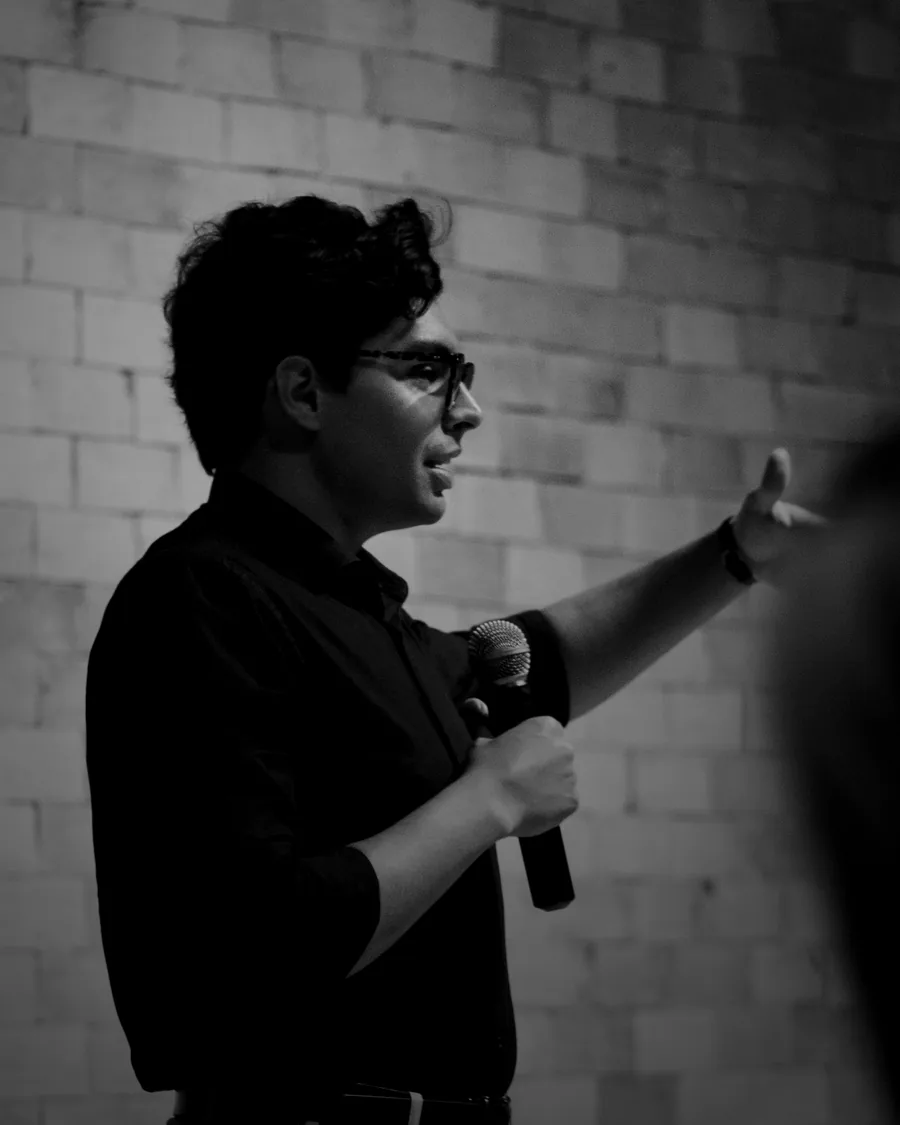 Lucas Quaranta de perfil, em preto e branco, falando ao microfone com a mão esquerda aberta no ar. Ao fundo, parede de tijolo aparente; na base do quadro, cabeças da plateia em contraluz.