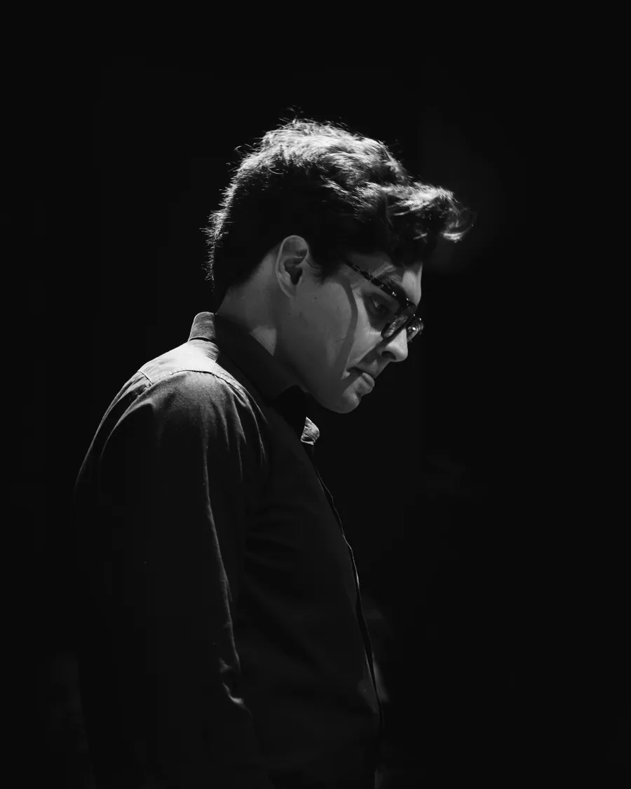 Retrato em preto e branco de Lucas de perfil, de cabeça baixa, recortado por uma luz lateral dura sobre fundo preto.